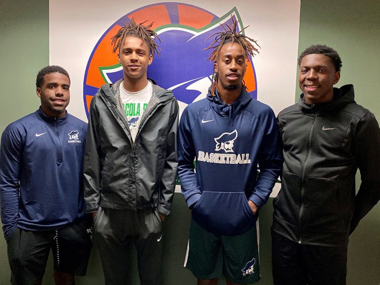 PSC Pirates basketball team members in athletic gear standing in front of school logo.