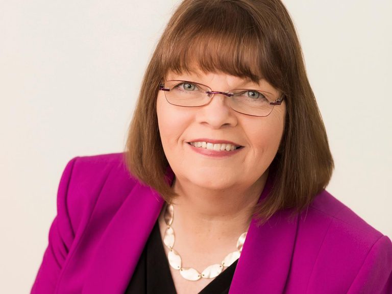 Senior woman in professional attire with glasses, smiling, representing college management change.