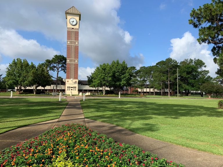 Prominent clock tower at Pensacola State College with lush green lawns and vibrant flower beds.