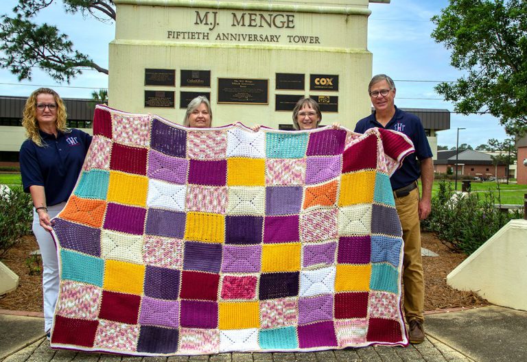 Group of volunteers holding colorful quilt in front of M.J. Menge Fifth Anniversary Tower.
