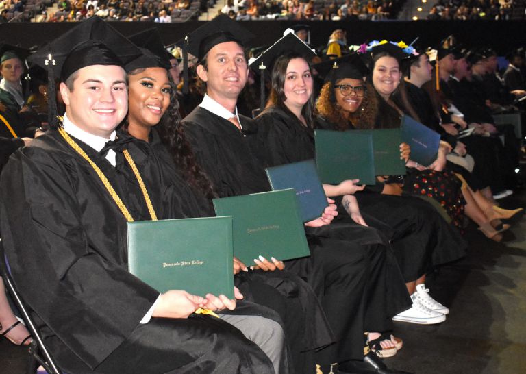 Group of diverse graduates in caps and gowns holding diplomas at Pensacola State graduation.