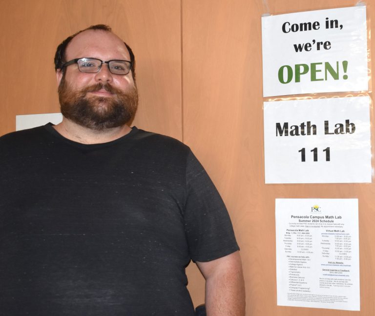 Former craftsman with a beard and glasses stands in front of a sign for Math Lab 111 at Pensacola St.